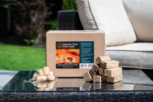 Box of Premier Pizza Oven Logs on glass table in garden with pizza oven logs and natural wood wool firelighters in the foreground