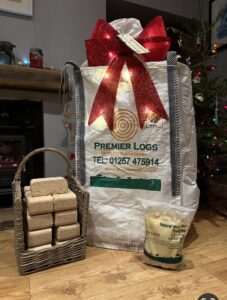 Premier Logs bag with red bow on, in front of Christmas tree and fire with Premier Logs Briquettes in basket and Premier Logs Natural Wood Wool Firelighters
